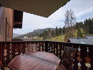 A wooden balcony with a table and chairs, overlooking a mountainous landscape with trees and buildings. - Apartment for 7, ski-in/ski-out, balcony (Morillon 1100)