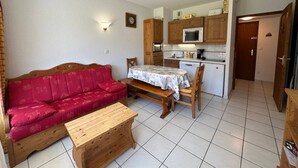 Ein kompakter Wohnbereich mit einem roten Sofa, einem Holztisch und einem Esstisch mit Stühlen. Eine Küchenzeile mit Holzschränken und einer Mikrowelle ist sichtbar.