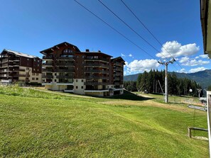 Un immeuble à plusieurs étages doté de balcons, situé sur une pente herbeuse avec des montagnes en arrière-plan.