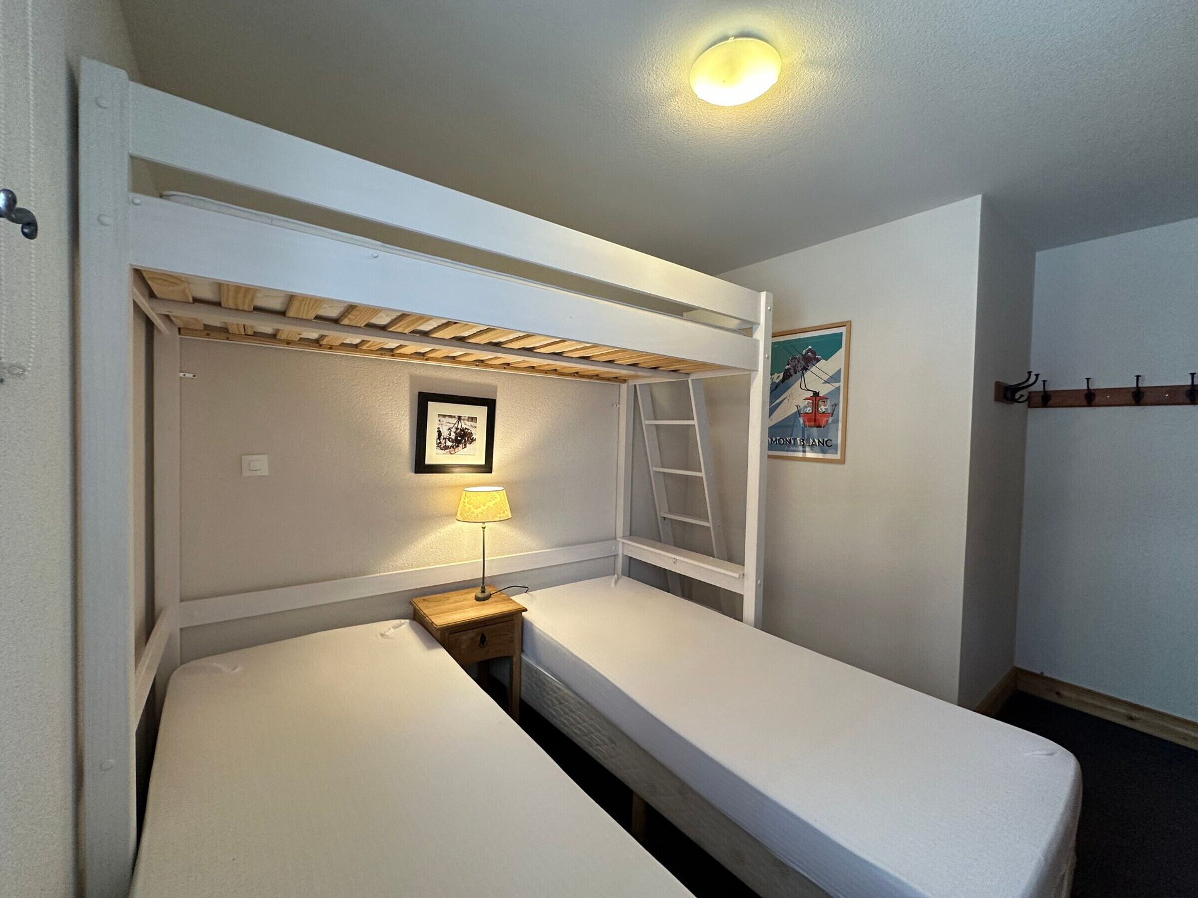 A bunk bed room with a ladder, a small bedside table with a lamp, a framed picture on the wall, and a coat rack.