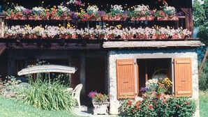 A two-story building with a balcony adorned with flowers, a small porch with a chair, and a stone wall. - Charming apartment in renovated farmhouse, sauna & terrace (Morillon Village)