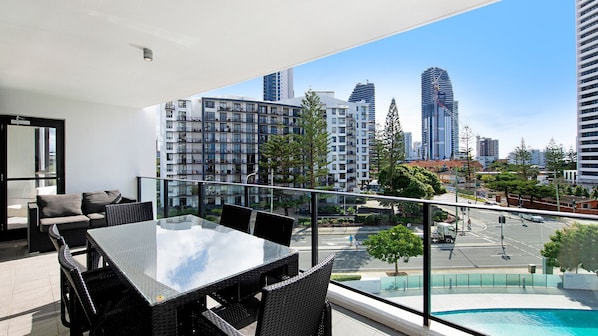 Un balcon doté d’une rambarde en verre, offrant une vue sur le paysage urbain, avec des bâtiments modernes et une piscine.