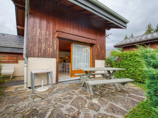 A rustic wooden building with a stone patio, a wooden door, and a table and chair set outside.