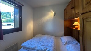 A bedroom with a bed, a window, a wall-mounted light, and wooden cabinets. - Charming 6-person apartment, balcony with mountain view, modern comfort, non-smoking (Morillon 1100)