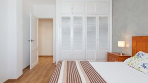 Ein Schlafzimmer mit Bett, Nachttisch, Lampe, einem Holz-Kopfteil, einem Streifen-Teppich und einem weißen Schrank mit Klappläden.