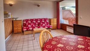 A small, cozy room with a wooden bed, a wooden chair, a sofa with a heart-patterned cover, a wooden dresser, and a window with a view of a wooden deck.