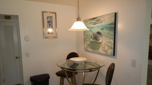 Uma pequena sala de jantar com mesa de vidro, duas cadeiras, um lixo e um quadro na parede.