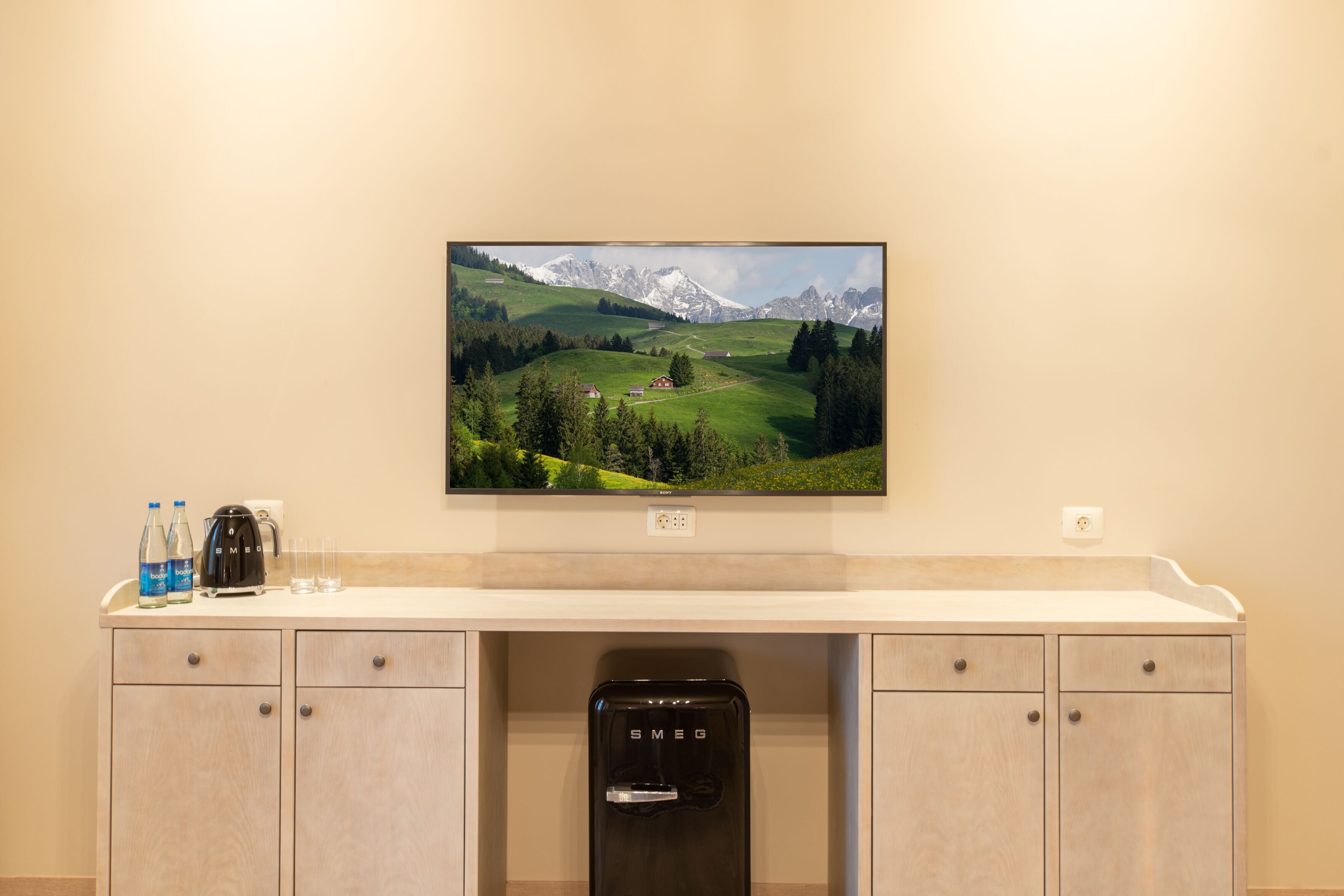 A hotel room with a large flat-screen TV mounted on the wall, a minibar with a water dispenser, and a wooden cabinet with drawers.