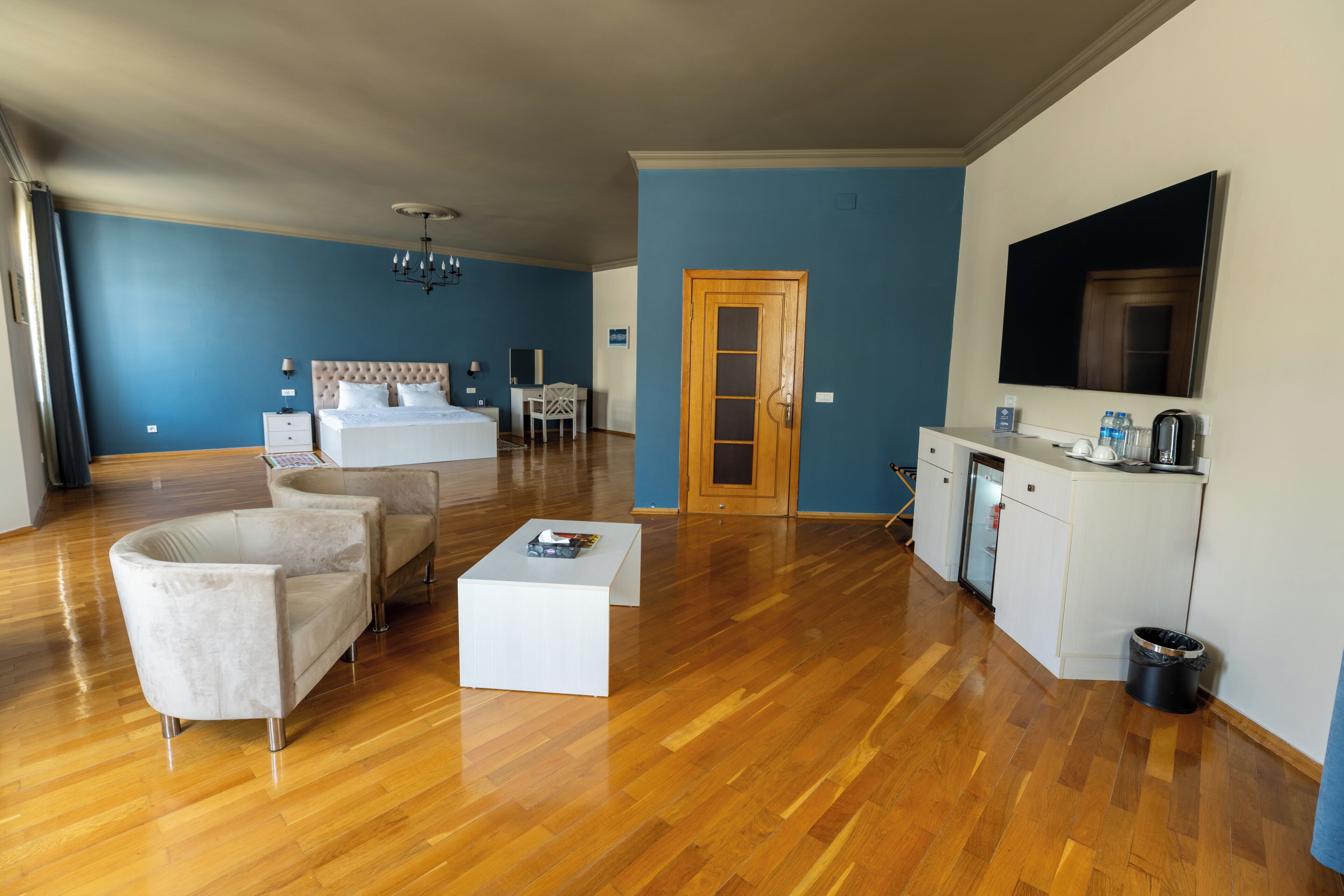 A hotel room with a bed, two armchairs, a coffee table, a desk, a TV, and a kitchenette.