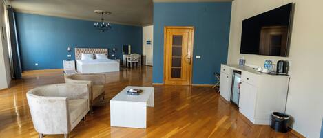 A hotel room with a bed, two armchairs, a coffee table, a desk, a TV, and a kitchenette.