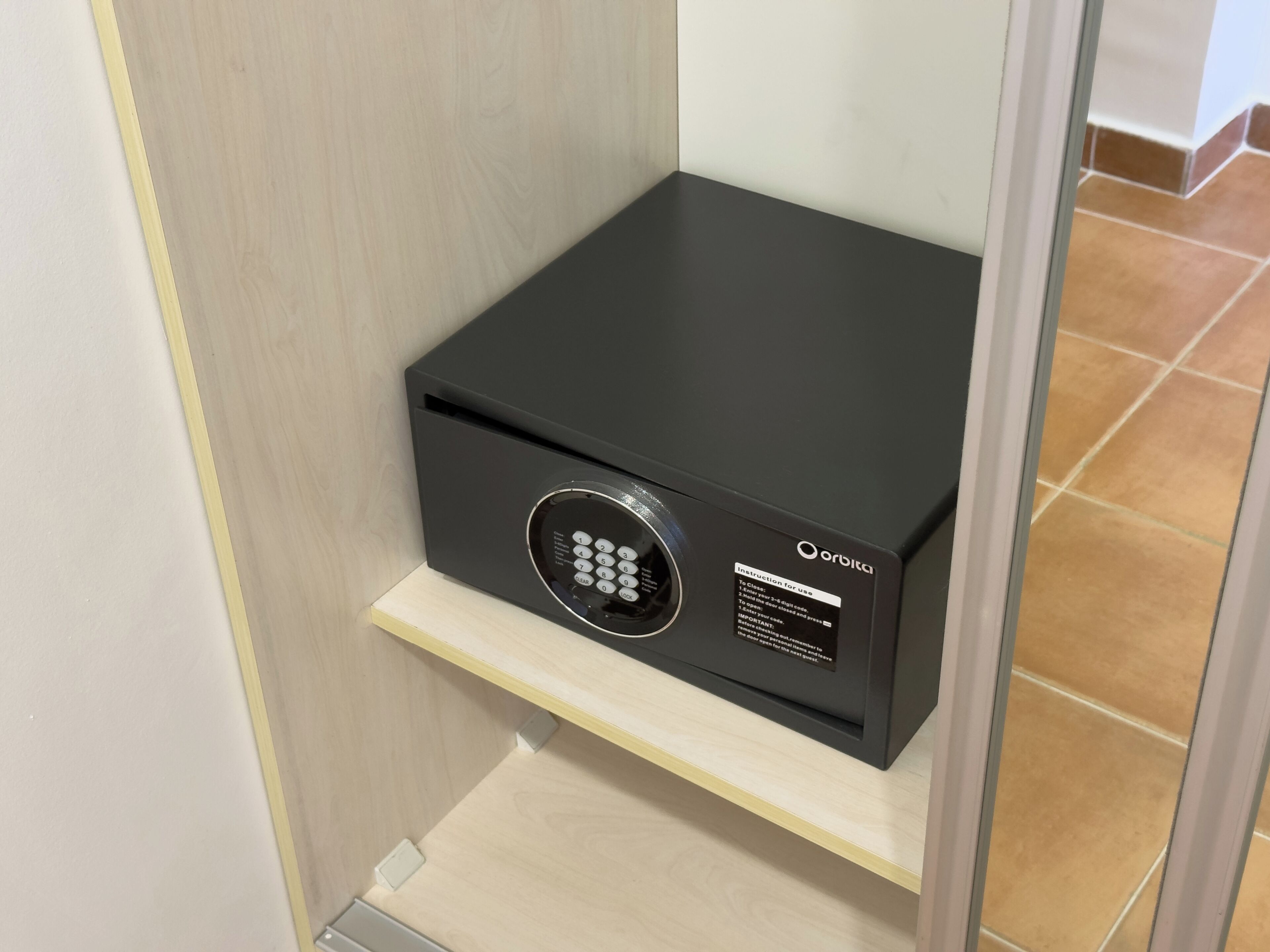 A black safe with a keypad, placed on a wooden shelf inside a room with tiled flooring.