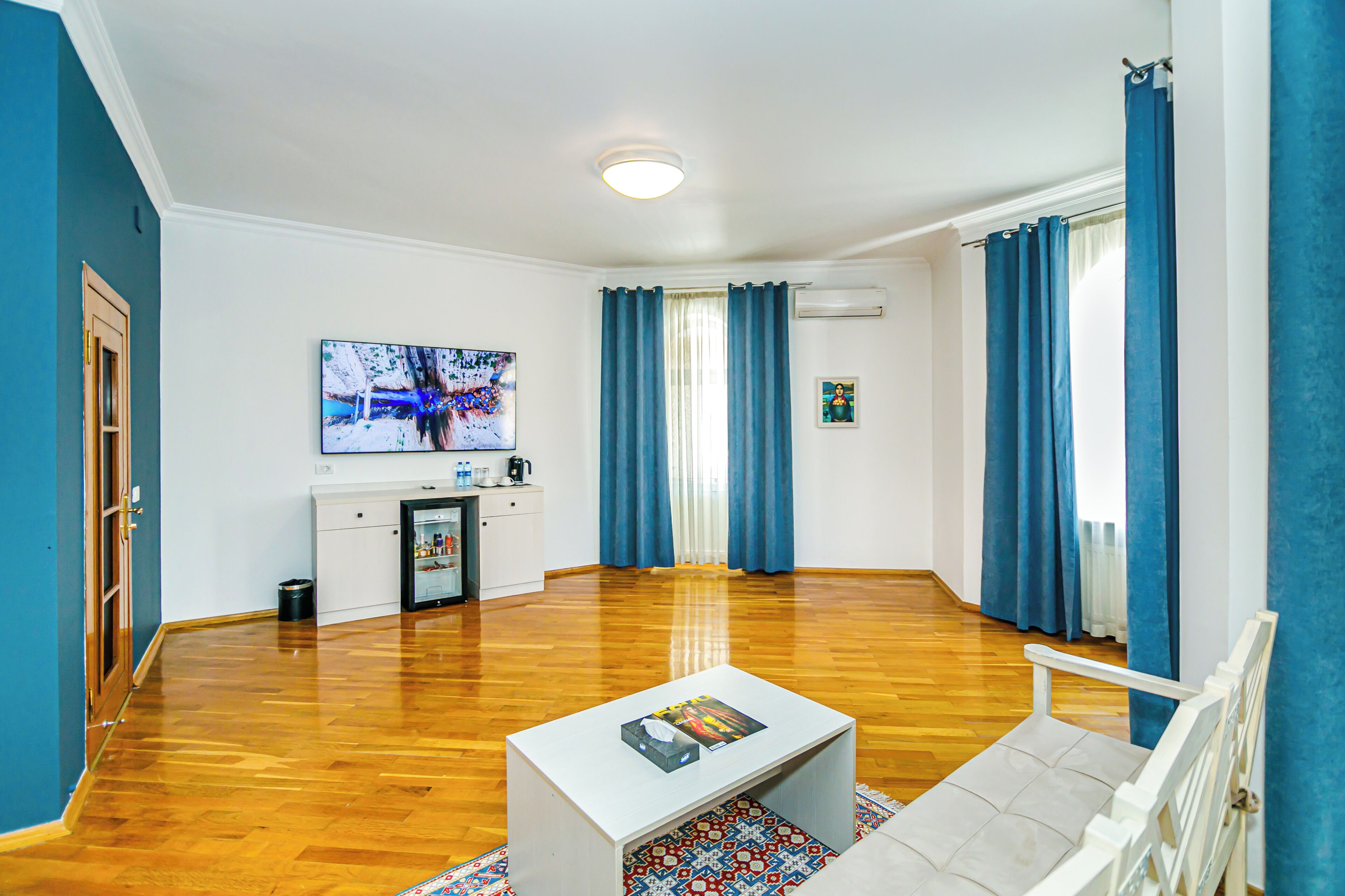 Una sala de estar con una mesa de centro blanca, un sofá, un televisor y una ventana con cortinas azules.