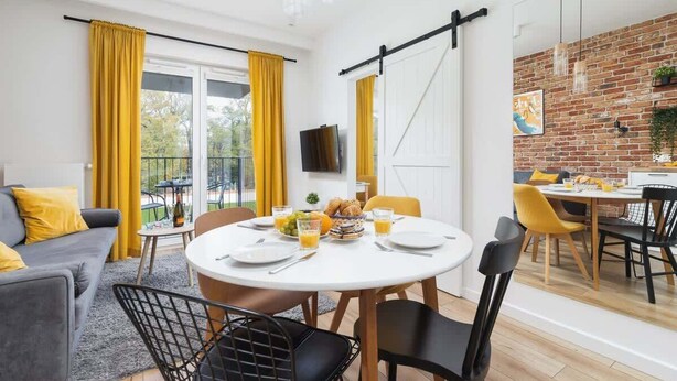 A modern dining area with a round table, yellow curtains, and a brick wall. - Stylish Apartment | Jacuzzi, Parking, Air Conditioning (Kraków)