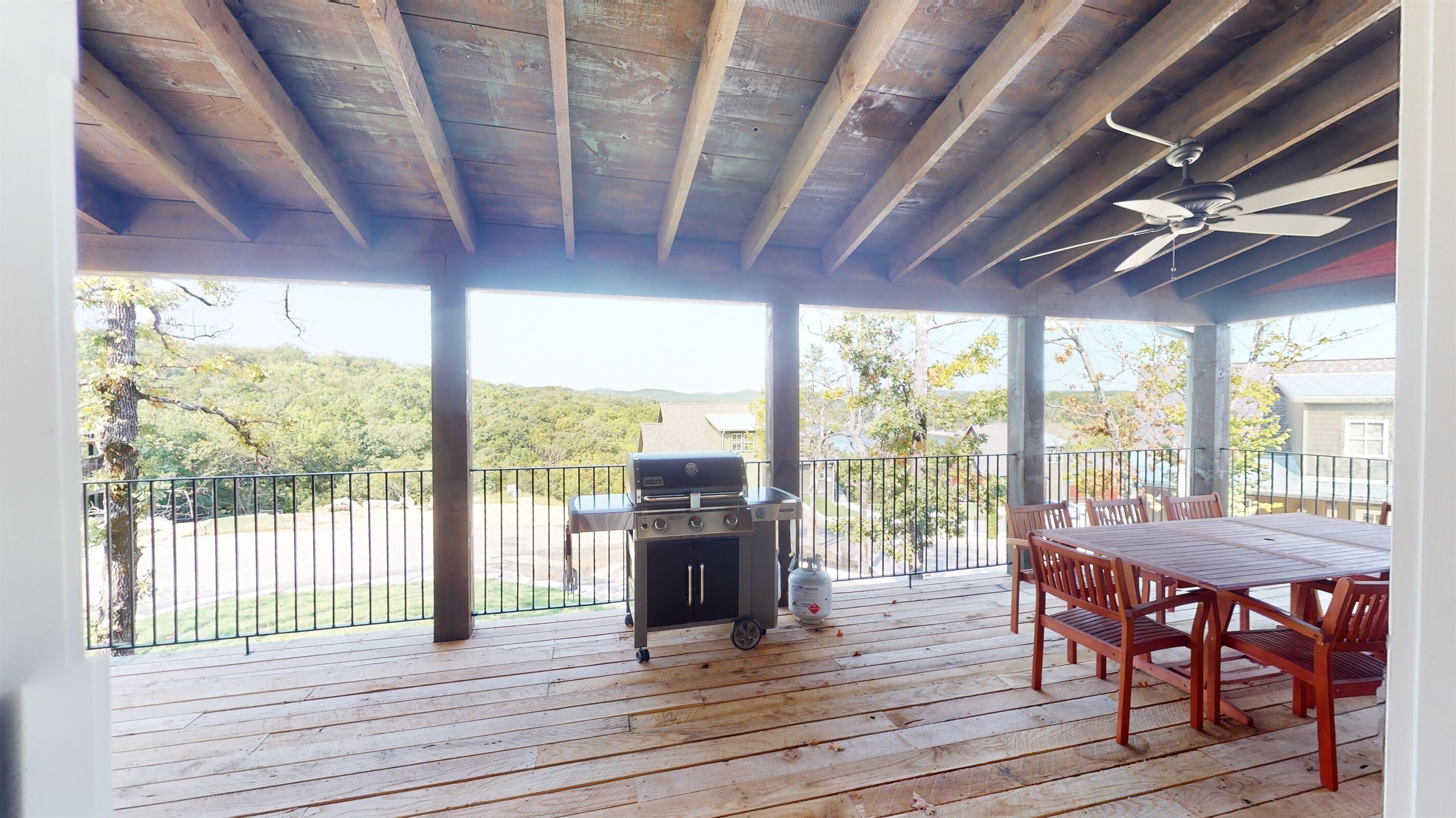 Een overdekt terras buiten, met een barbecue, houten tafel en stoelen.