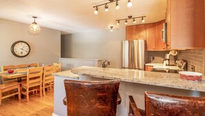 A modern kitchen with wooden cabinets, a stainless steel refrigerator, a granite countertop, and a dining area with wooden chairs and a table. - Nice Updated Condo. Hot Tub. Access to Lake. (Frisco)