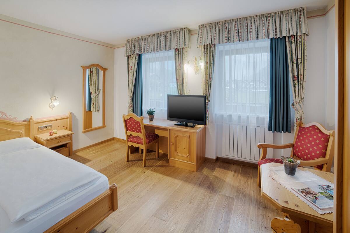 Une chambre d’hôtel avec un lit, un bureau avec un ordinateur, une chaise, une petite table avec une plante et une fenêtre avec des rideaux.