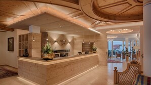 A reception area with a wooden reception desk, pendant lighting, and a view into a dining area with chairs and tables. - Alpenblick Hotel (Valle Aurina)