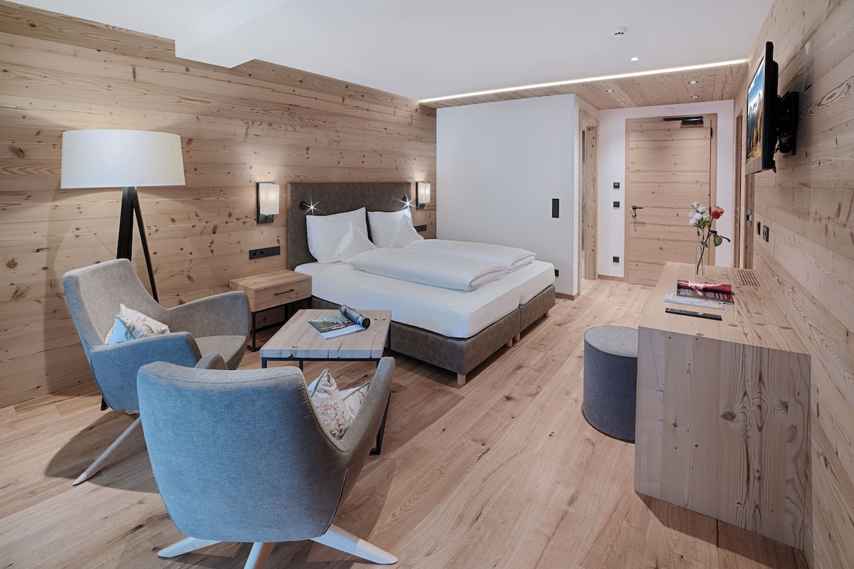 Une chambre moderne avec un mur décoré de panneaux en bois, un lit recouvert de linge de maison blanc, un fauteuil gris, une table de chevet avec une lampe, et une petite table sur laquelle sont posés des livres et un vase de fleurs.