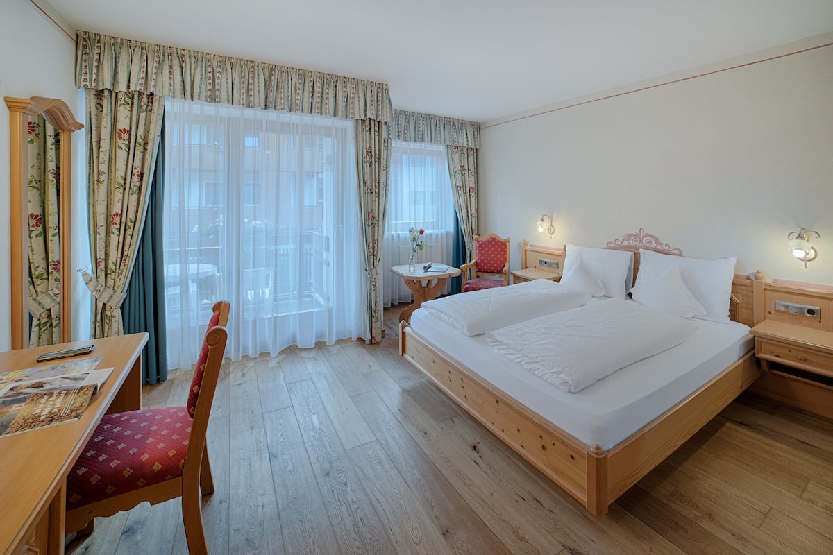 Une chambre d’hôtel avec un lit en bois, un bureau en bois, une chaise, une petite table avec un vase et des rideaux.