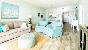 A living room with a beige sofa, a blue armchair, and a round ottoman. There is a painting of sailboats on the wall and a chandelier in the dining area.