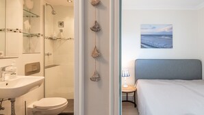 A hotel bathroom with a sink, toilet, and shower. There is a bed with a headboard and a lamp on a side table. - Beautiful 2-room vacation apartment in city location on approx. 50 m² for up to 2 persons. (Westerland)