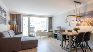 A modern living room with a sofa, dining table, and chairs. There is a window with curtains, a radiator, and a television. - Beautiful 2-room vacation apartment in city location on approx. 50 m² for up to 2 persons. (Westerland)