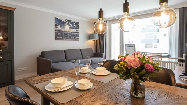 A dining area with a wooden table set for two, a vase with flowers, and a cityscape painting on the wall. - Beautiful 2-room vacation apartment in city location on approx. 50 m² for up to 2 persons. (Westerland)