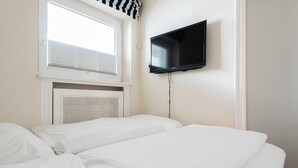 A hotel room with two beds, a TV mounted on the wall, and a window with blinds. - Beautiful 2-room vacation apartment in city location on approx. 50 m² for up to 2 persons. (Westerland)