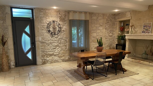 A dining area with a stone wall, a wooden table, and chairs. There is a black door with a window, a wall clock, and a fireplace with a mantel. - mas (Tarascon)