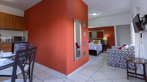 Habitación de hotel con cocina, una mesa de comedor con sillas, una cama con mesita de noche y un televisor montado en la pared.