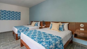 Habitación de hotel con dos camas, cada una con cubrecama de estilo tropical en azul y blanco, cabeceros de madera y mesita de noche con lámpara.