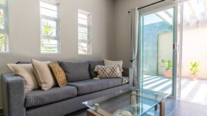 Una sala de estar moderna con un sofá gris, una mesa de centro de vidrio y ventanas amplias con cortinas.