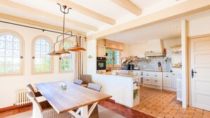 A spacious kitchen with a central island, large windows, and a dining area with a wooden table and wicker chairs.