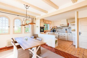 A spacious kitchen with a central island, large windows, and a dining area with a wooden table and wicker chairs.
