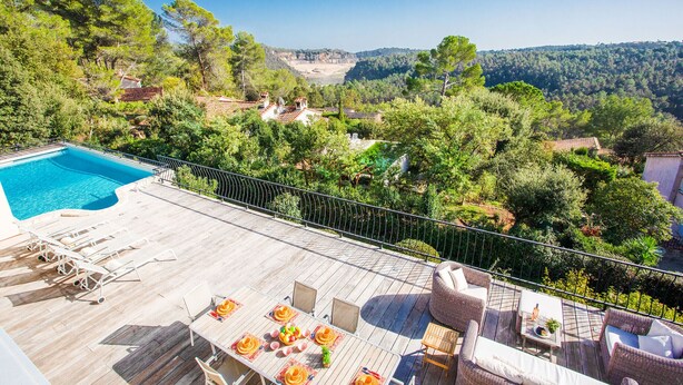 A poolside area with lounge chairs, a dining table, and a view of a forested valley.
