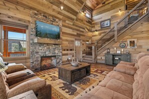 A log cabin interior with a stone fireplace, a large TV, a staircase, and a cozy seating area.