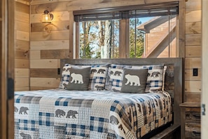 Ein Schlafzimmer in einer Holzhütte mit einem Bett, einem Nachttisch, einem Fenster mit Blick auf Bäume und einer Wandleuchte.