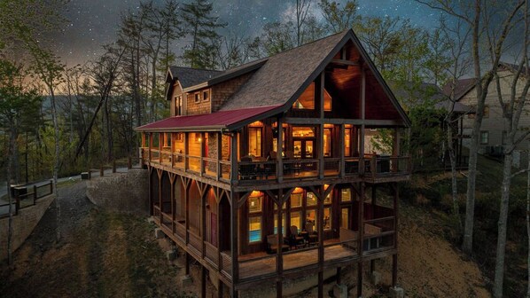 Eine zweistöckige Blockhütte mit Balkon, umgeben von Bäumen und einem sternenübersäten Nachthimmel.
