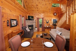 A cozy cabin interior with a fireplace, wooden walls, a sofa, and a dining table set for two.