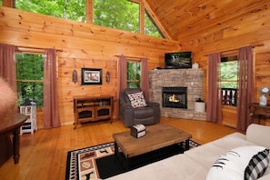 A cozy cabin interior with wooden walls, a stone fireplace, a leather sofa, and a large window overlooking greenery.