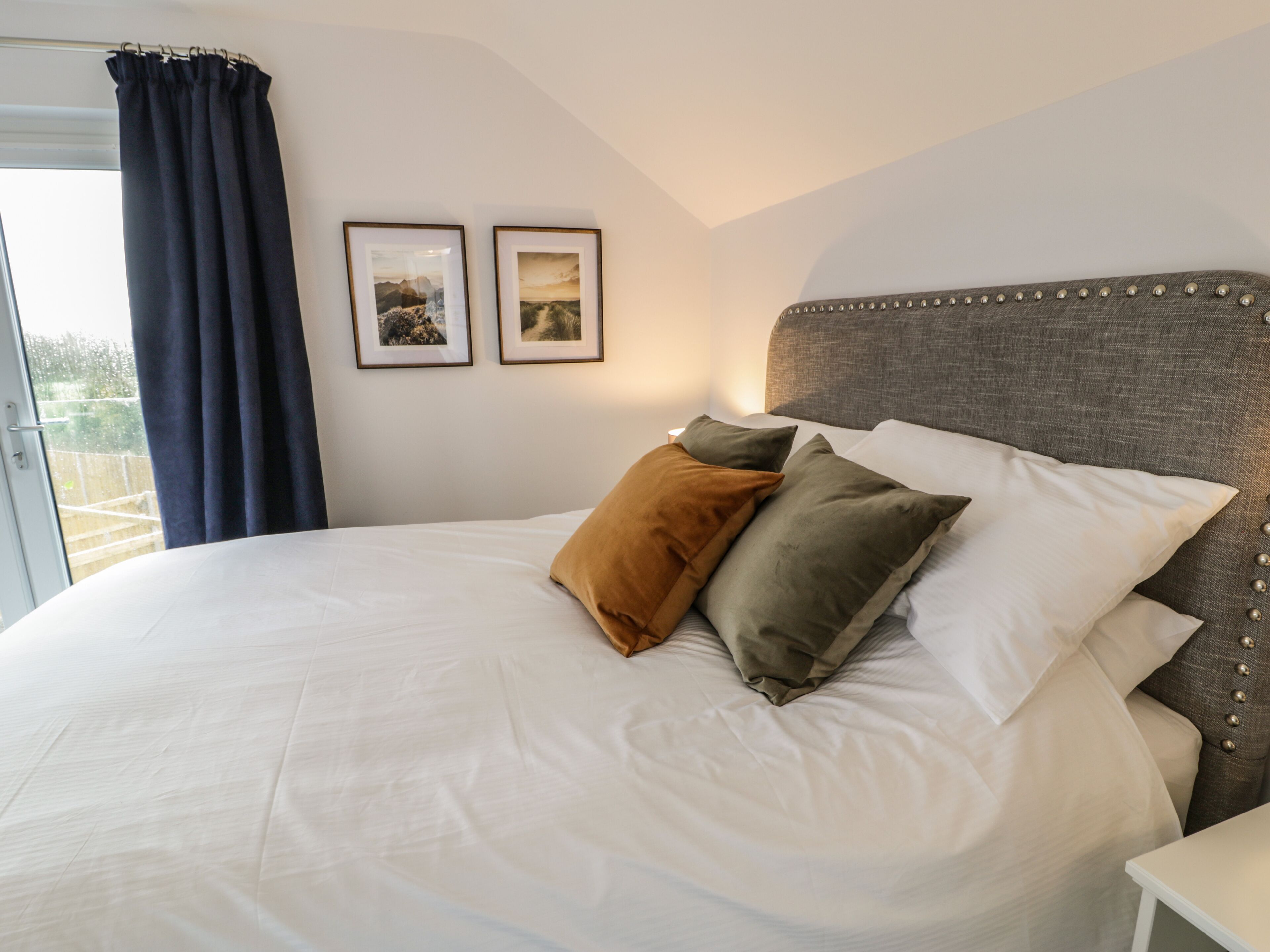 Een net opgemaakt bed met witte lakens en meerdere kussens, een grijs bekleed hoofdbord, en twee ingelijste foto's aan de muur.