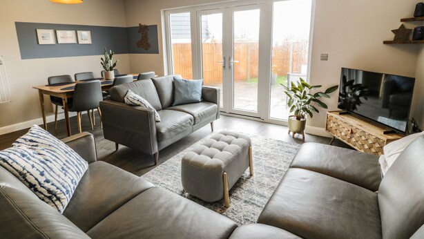 Ein modernes Wohnzimmer mit grauem Sofa, Hocker und einem Essbereich mit Tisch und Stühlen.