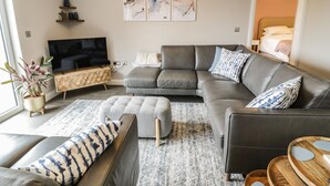 Ein modernes Wohnzimmer mit einem grauen Ecksofa, einem kleinen Hocker, einer Holz-TV-Bank und einem großen Fenster.