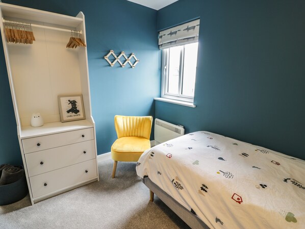A bedroom with a bed, a chair, a dresser, a radiator, and a window with a curtain. - Can y Mor (Tyn-y-Gongl)