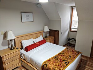 A bedroom with a wooden bed, two bedside tables, a nightstand, a red throw pillow, a wall-mounted lamp, a framed sign, and a window with blinds.