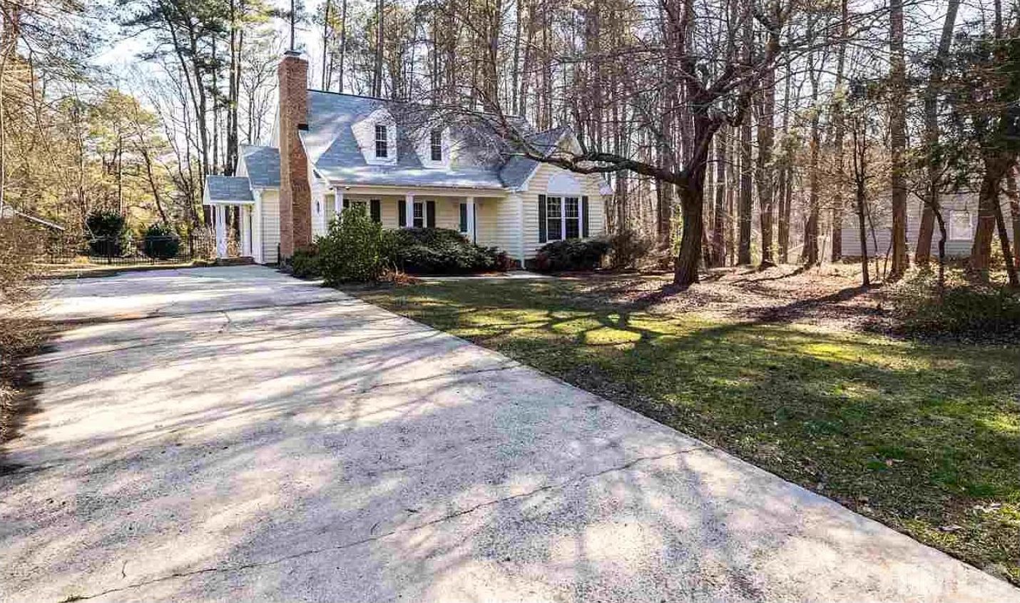 A two-story house with a front yard, a driveway, and a wooded area.