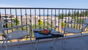 Un balcón con dos sillas blancas y una mesa, un plato de fruta, y vistas a edificios y árboles.