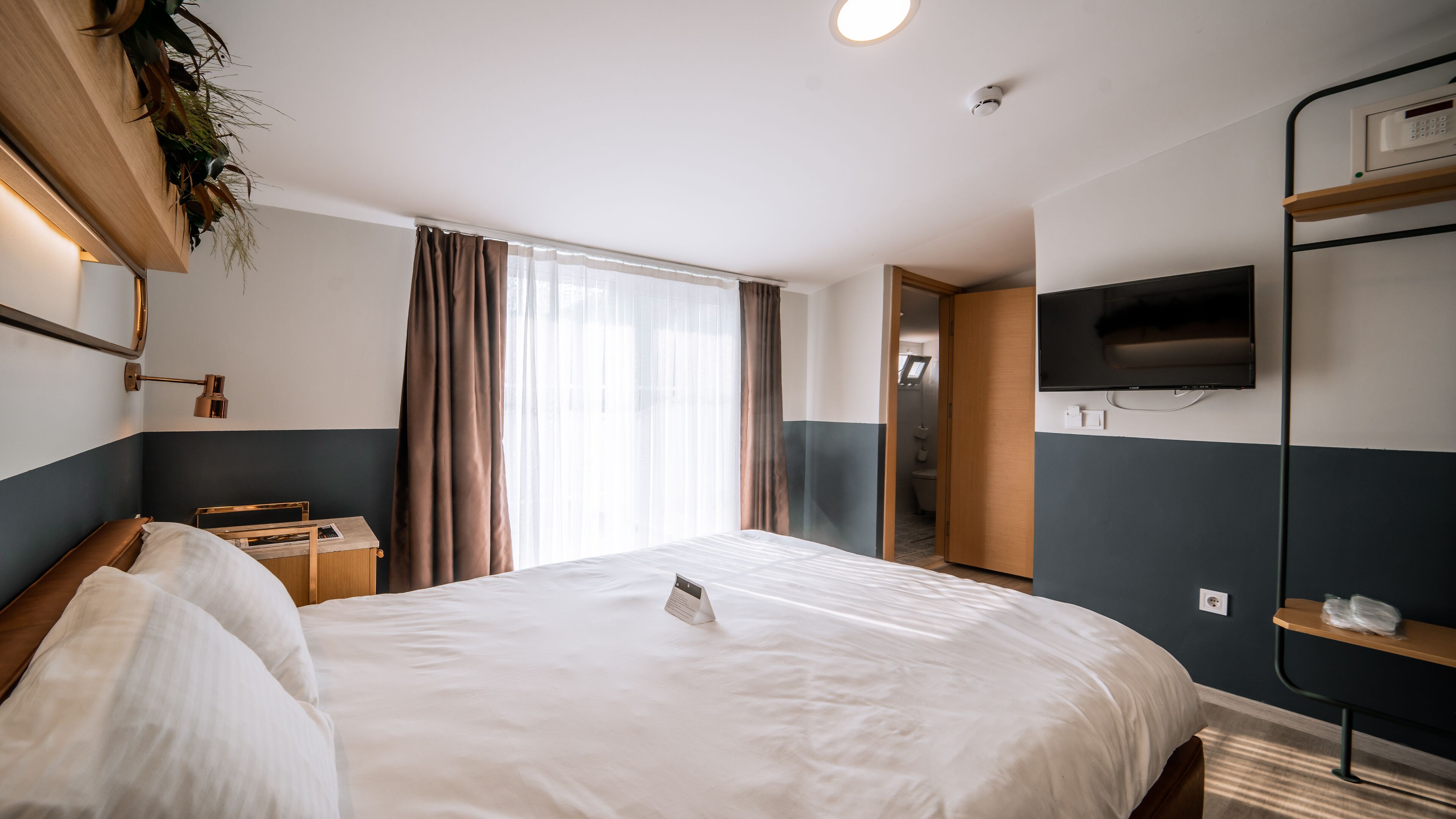 A hotel room with a bed, bedside table, lamp, TV, and a view of the bathroom.