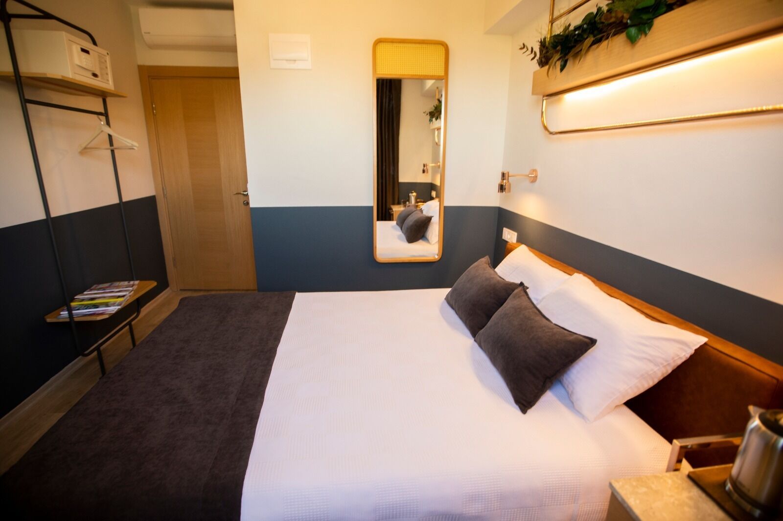 A hotel room with a bed, a bedside table with a lamp, a mirror, and a shelf with a plant.