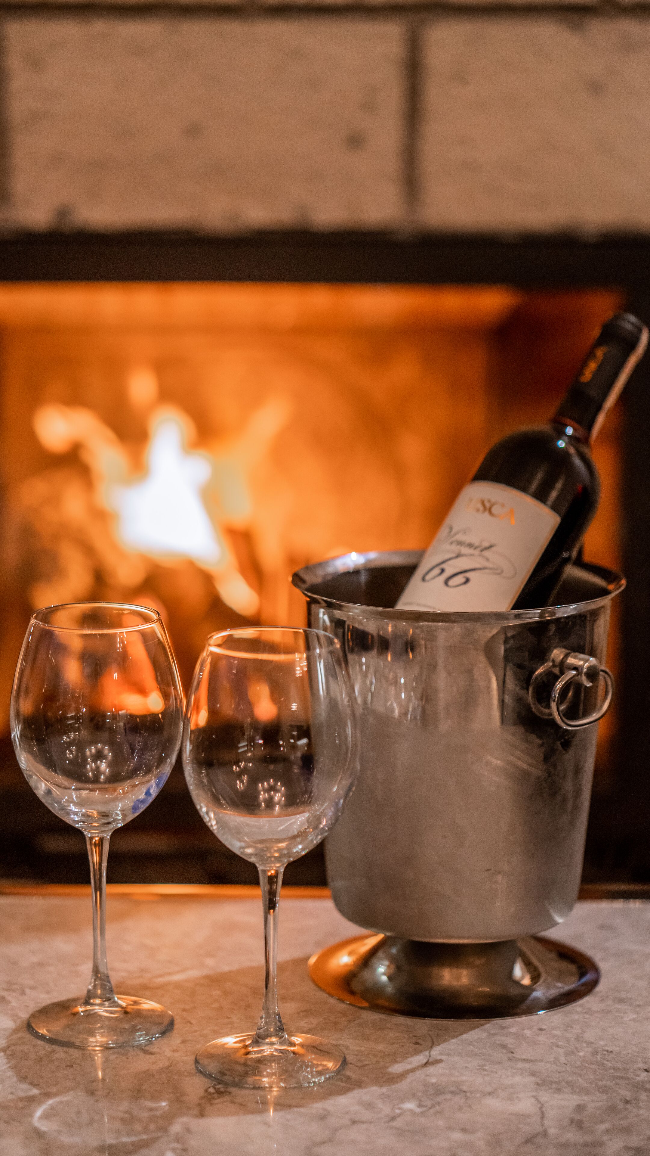 Duas taças de vinho e uma garrafa de vinho em um balde de metal sobre uma superfície de mármore, com uma lareira ao fundo.